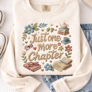 May include: Cream-colored sweatshirt with the phrase "Just One More Chapter" in a textured, beige font. The design includes books, flowers, a teapot, and glasses, creating a cozy, book-themed aesthetic. The sweatshirt has a crew neck and long sleeves.