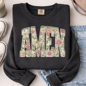 Puede incluir: Una sudadera negra con la palabra "AMEN" en un diseño floral. Las letras están rellenas con un patrón de flores rosas, blancas y amarillas sobre un fondo verde claro. La sudadera está colocada sobre una superficie blanca.