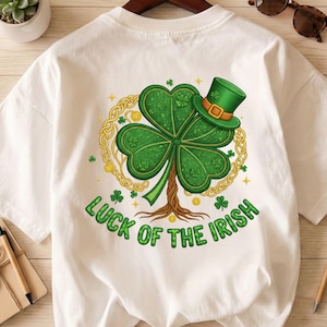 May include: White t-shirt with a green shamrock design, a green top hat, and gold detailing. The text "LUCK OF THE IRISH" is printed in green. The graphic is centrally placed on the shirt.