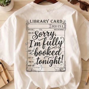 May include: White t-shirt featuring a library card design with the text "Sorry, I'm fully booked tonight!". The design includes dates and book illustrations, perfect for book lovers.
