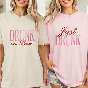 Op de afbeelding: Twee T-shirts, één beige en één roze, met de woorden "DRUNK in Love" en "Just DRUNK" gedrukt in roze en rode letters. De shirts hebben korte mouwen en een relaxte pasvorm.
