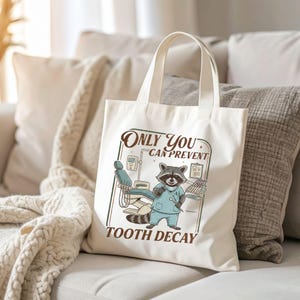 Op de afbeelding: Cr&egrave;mekleurige draagtas met een cartoon wasbeer als tandarts. De wasbeer staat voor tandheelkundige apparatuur. De tekst op de tas luidt: "Only You Can Prevent Tooth Decay".