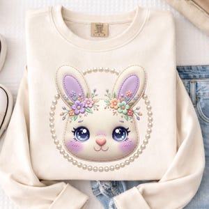 Peut inclure: Sweat-shirt crème avec un visage de lapin de dessin animé. Le lapin a de grands yeux bleus, un nez rose et des accents violets et floraux, entouré d'un cercle de perles. Le sweat-shirt est associé à un jean bleu.