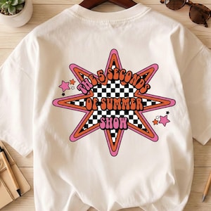May include: Cream-colored t-shirt featuring a retro design. The graphic includes a star shape with pink, orange, and black accents, and the text "THE 5 SECONDS OF SUMMER SHOW". Small pink stars are scattered around the main design. A casual, unisex tee.