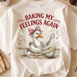 May include: White t-shirt with a cartoon goose wearing a pink bow and apron, whisking batter in a bowl. The text "BAKING MY FEELINGS AGAIN" is arched above the goose. The design includes baking-related elements.