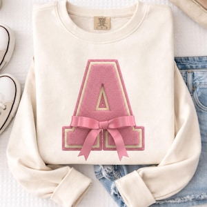 May include: Cream-colored sweatshirt featuring a large, pink letter "A" with a light yellow border and a pink bow. The letter has a textured appearance. The sweatshirt is folded on a white surface, with a pair of white sneakers and denim jeans visible.