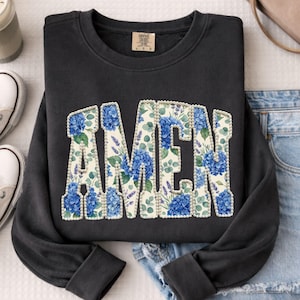 Op de afbeelding: Zwart sweatshirt met het woord "AMEN" in een bloemenontwerp. De letters zijn gevuld met blauwe hortensia's, lavendel en groen, omlijnd met een parelachtige rand. Het sweatshirt ligt plat op een witte ondergrond.