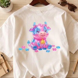 May include: White t-shirt featuring a cartoon cow design. The cow is pink and blue with a gold bell around its neck. Scattered around the cow are pink and blue shapes. The shirt is hanging on a wooden hanger.