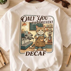 Op de afbeelding: Wit t-shirt met een cartoon kat barista ontwerp. De afbeelding bevat de tekst "ONLY YOU CAN PREVENT DECAF." De kat draagt een schort en houdt een kopje koffie vast, in een koffiebar met een koffiemachine en molen.