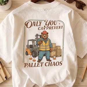 Op de afbeelding: Wit T-shirt met een cartoonbeer met een helm en veiligheidsvest, die een vorkheftruck bedient. De tekst luidt: "ONLY YOU CAN PREVENT PALLET CHAOS."