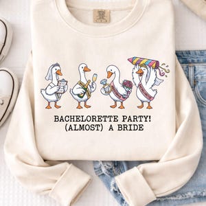 May include: Cream-colored sweatshirt featuring a whimsical illustration of geese dressed for a bachelorette party. The geese are holding items like a clipboard, champagne, a ring, and a firework. Text at the bottom reads "Bachelorette Party! (Almost) A Bride."