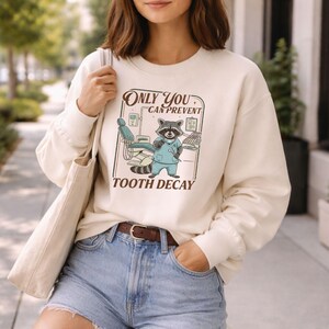 Op de afbeelding: Cr&egrave;mekleurige sweatshirt met een afbeelding van een wasbeer-tandarts en de tekst "Only You Can Prevent Tooth Decay". De wasbeer draagt een blauw uniform en staat voor een tandartsstoel. Het ontwerp is in vintage stijl.