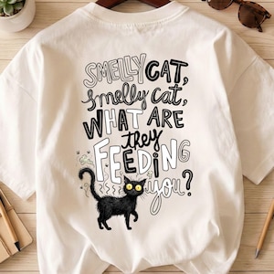 Puede incluir: Camiseta blanca con un gato negro de dibujos animados con ojos amarillos y el texto "Smelly Cat, Smelly Cat, What Are They Feeding You?" en una fuente lúdica. El gato tiene una expresión de sorpresa.