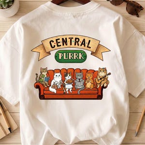May include: White t-shirt featuring a cartoon illustration of six cats on a red couch. The cats are depicted in various poses, with one playing a guitar. The text "CENTRAL PURRK" is above the couch in a banner.
