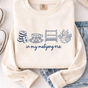 May include: Cream-colored sweatshirt with a blue graphic of Mahjong tiles, a teacup, a rack of tiles, and a dragon. The text "in my mahjong era" is written in blue script below the graphic.