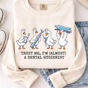 Op de afbeelding: Crèmekleurige sweatshirt met een grillig ontwerp van vier cartoon ganzen verkleed als mondhygiënisten, met tandheelkundige instrumenten. De tekst "TRUST ME, I'M (ALMOST) A DENTAL HYGIENIST" staat eronder. De sweatshirt wordt gecombineerd met een blauwe jeans.