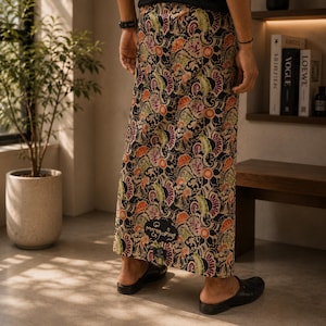 May include: A floor-length sarong featuring a dense, intricate pattern of orange, green, and purple floral and paisley motifs on a black background. The fabric wraps around the lower body, paired with a black t-shirt and black slip-on shoes.