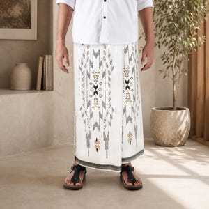 May include: A white sarong featuring a geometric pattern in shades of black, gray, and gold. The fabric is wrapped around the waist, extending to the lower legs, and displays intricate symmetrical motifs centered on the front panel of the garment.