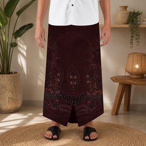 May include: A floor-length, deep burgundy sarong featuring intricate, dark-toned floral and swirling patterns throughout the fabric. The garment is worn with a crisp white button-down shirt and black open-toe sandals, creating a clean silhouette.