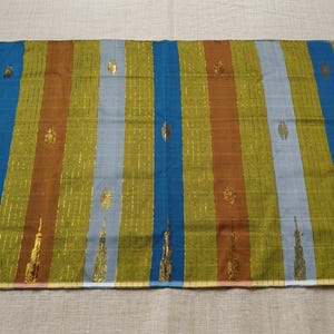 Puede incluir: Un tejido rectangular con rayas verticales en tonos verde oliva, marrón, azul claro y azul oscuro. La tela está decorada con finos hilos metálicos dorados verticales y motivos bordados en oro que se repiten.