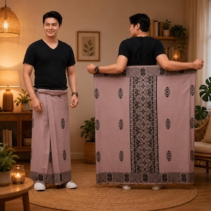 May include: A mauve sarong featuring a black geometric pattern along the hem and a central vertical panel with intricate black detailing. The fabric is worn wrapped around the waist, paired with a plain black v-neck t-shirt and white trainers.