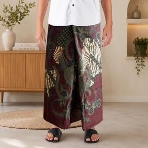 Könnte beinhalten: Ein langer, weit geschnittener Rock mit einer tiefburgunderroten Grundfarbe und einem detaillierten Batik-Muster in Dunkelgrün, Creme und Schwarz. Der Stoff zeigt ein markantes Vogelmotiv, das von komplexen Blumen- und Rankenmustern umgeben ist.