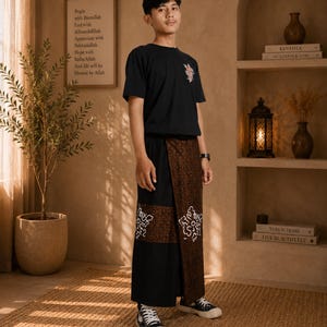 Könnte beinhalten: Ein schwarzes kurzärmeliges T-Shirt mit einer kleinen Blumen-Grafik auf der Brust, kombiniert mit einem schwarz-braunen Wickelrock mit komplexen Batik-Mustern und weißen dekorativen Motiven am unteren Saum, getragen mit schwarzen Turnschuhen.