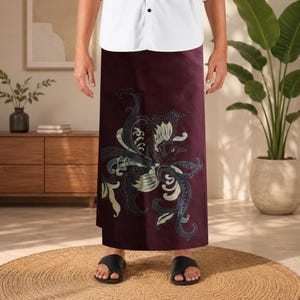 May include: A deep burgundy sarong featuring a large, intricate batik-style phoenix motif in shades of cream, teal, and dark blue. The fabric is wrapped around the waist, paired with a plain white button-down shirt and black slide sandals.