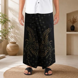 Puede incluir: Un pareo negro con un patrón de batik en tono dorado que presenta motivos simétricos en forma de alas y un estampado geométrico de rombos. La prenda se combina con una camisa blanca de botones y sandalias negras, creando una silueta recta.
