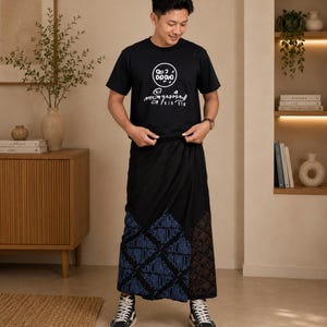 May include: A black short-sleeved t-shirt featuring a white circular graphic with text above a script-style inscription, paired with a black wrap-around skirt that has a blue geometric pattern on one side and a dark brown floral print on the other.