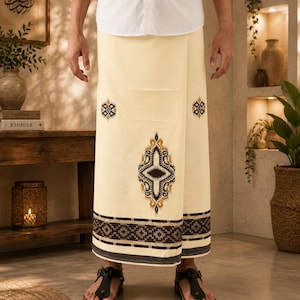 May include: A cream-colored sarong featuring a central dark brown and gold geometric motif, smaller matching side accents, and a wide horizontal border at the hem with intricate dark brown and white patterned stripes and solid dark bands.