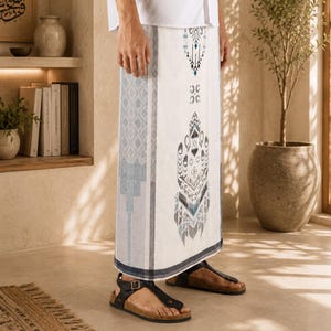 May include: A white sarong featuring a vertical panel with intricate dark blue and grey geometric patterns, worn with black thong-style sandals. The fabric displays a central decorative motif and side borders with repeating diamond shapes.