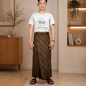 Puede incluir: Una camiseta de manga corta gris claro con un gráfico circular negro y texto estilizado, junto a un pareo de batik marrón con un patrón diagonal repetitivo. El conjunto se completa con sandalias negras sencillas de punta abierta.