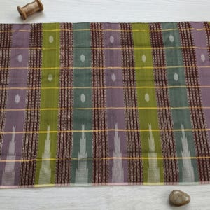 Puede incluir: Un tejido rectangular con un patrón de rayas verticales en tonos verde oliva, granate oscuro y morado apagado. La tela incluye hilos metálicos dorados horizontales finos y pequeños motivos repetitivos en forma de diamante blanco.