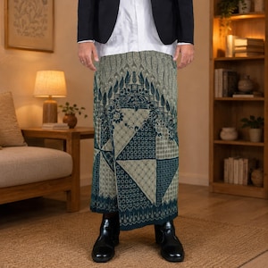 May include: A long, wrap-style skirt featuring a dark teal and beige batik pattern with geometric and floral motifs. The skirt is worn with a white button-down shirt, a black blazer, and black leather ankle boots, creating a layered ensemble.