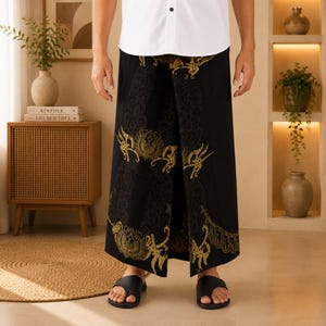 Può includere: Un sarong nero caratterizzato da un intricato motivo color oro con disegni simmetrici. Il capo è indossato con una camicia bianca abbottonata e sandali infradito neri. Il tessuto presenta un design ripetitivo che copre la parte anteriore.