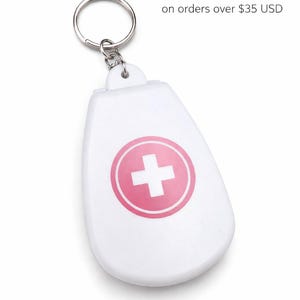 Könnte beinhalten: Weißer Schlüsselanhänger mit einem rosa Kreuzsymbol in einem rosa Kreis. Der Schlüsselanhänger hat einen silbernen Ring und eine weiße, abgerundete rechteckige Form. Der Text "Free Shipping" ist sichtbar.