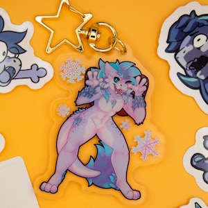 Puede incluir: Un llavero acrílico transparente con un personaje de dibujos animados con pelaje azul y morado, detalles de copos de nieve y un cierre dorado en forma de estrella. El personaje está en una pose juguetona, rodeado de pegatinas sobre un fondo amarillo.