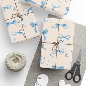 Könnte beinhalten: Geschenke mit hellblauem Schleifen- und Bandmuster auf cremefarbenem Papier verpackt. Jedes Paket ist mit Bindfaden und einem weißen Anhänger verschnürt. Eine Garnrolle, eine Schere und leere Anhänger sind ebenfalls zu sehen.