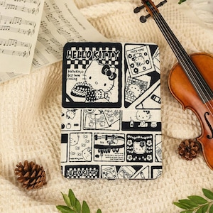 May include: A rectangular e-reader case featuring a black and white comic-style print of Hello Kitty. The design includes multiple panels with illustrations of the character, the text HELLO KITTY, and the phrase Hello Kitty's best home cooking.