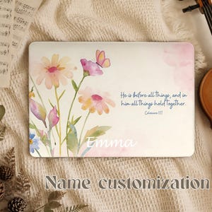 Könnte beinhalten: Eine rechteckige weiße Platte mit einer Aquarellillustration von Wildblumen in Rosa-, Gelb- und Blautönen sowie einem kleinen Schmetterling. Der Text lautet: "He is before all things, and in him all things hold together. Colossians 1:17" und "Emma."
