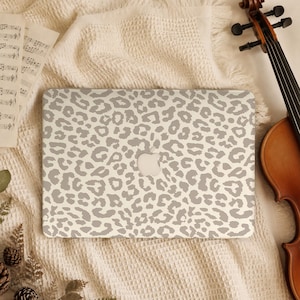 Op de afbeelding: Een laptop met een lichtgrijze en witte luipaardprint skin op de deksel. De laptop heeft een gecentreerd zilveren appel-logo. Het apparaat ligt op een textuurrijke crèmekleurige stof naast bladmuziek en een viool.