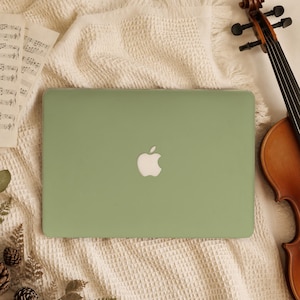 May include: A sage green laptop case with a centered white Apple logo, positioned on a textured cream-colored fabric next to a wooden violin neck and a sheet of musical notation paper with printed notes and staff lines.