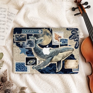 Op de afbeelding: Een laptopsticker met een collage van donkerblauwe en crèmekleurige elementen, waaronder een grote walvis, een volle maan, postzegels, een schelp en de tekst OCEAN BLVD. Het ontwerp bevat een centrale uitsparing voor het Apple-logo.