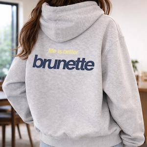 Peut inclure: Un sweat à capuche gris avec le texte "life is better brunette" en jaune et bleu marine. Le sweat à capuche a une capuche et des manches longues. La personne porte un jean bleu.