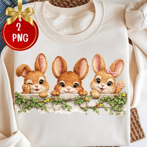 May include: Cream-colored sweatshirt featuring three cartoon bunnies peeking over a stone wall adorned with greenery and flowers. The bunnies have brown fur and pink accents, with one winking and another wearing a pink bow. The image includes a gold ribbon with the text "2 PNG".
