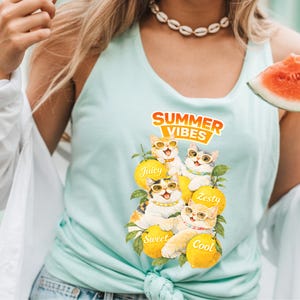 May include: A light blue tank top with a graphic of four cats wearing sunglasses, sitting on lemons. The text "Summer Vibes" is above the cats, with the words "Juicy", "Zesty", "Sweet", and "Cool" on the lemons.