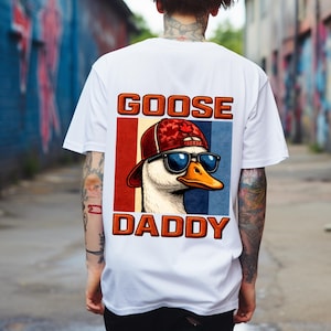 Pode incluir: Camiseta branca com um gráfico de um ganso usando óculos de sol e um chapéu. O texto "GOOSE DADDY" é exibido acima e abaixo da imagem do ganso em letras laranja.