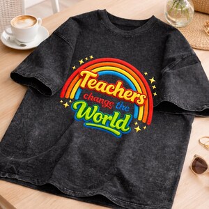 Op de afbeelding: Donkergrijs t-shirt met een regenboogontwerp en de tekst "Teachers change the World". Het ontwerp heeft een regenboogboog met gele en blauwe woorden. Het shirt is gemaakt van een zacht, comfortabel materiaal.