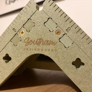 以下が含まれることがあります： エッチングされたマーキングと「Southam DESIGNWORKS」のテキストが付いた、明るい緑色の直角測定ツール。ツールには一連の切り抜かれた形状があり、金色のネジで固定されています。マーキングはセンチメートルです。